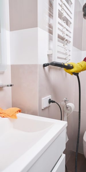 Cropped photo of man in rubber glove steam-cleaning joint between bathroom ceramic tiles