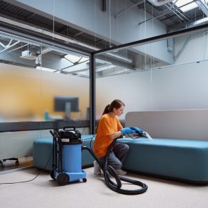 Hard-working female janitor squatting beside the sofa while carefully vacuum-cleaning the upholstery