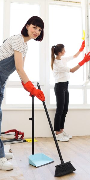 Red haired girl in overalls sweeping floors in house with broom and dustpan, and another female in casual clothes wiping the window. Young beautiful women cleaning up new apartment after renovation.